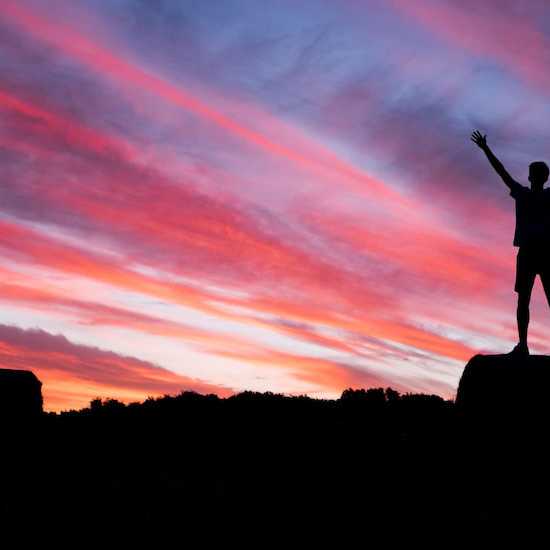 young person standing on a rock with their arms up looking into the sunset. life after cancer image