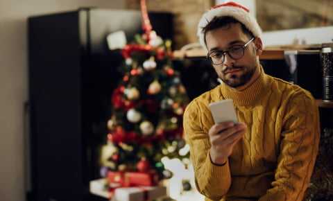 young person wearing a santa hat and sitting in front of a christmas tree while looking at their phone