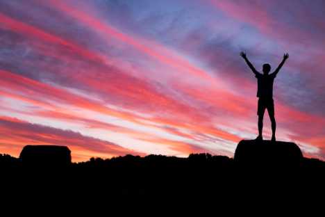 young person standing on a rock with their arms up looking into the sunset. life after cancer image