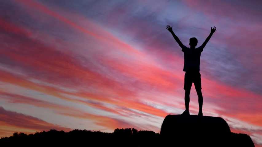 young person standing on a rock with their arms up looking into the sunset. life after cancer image