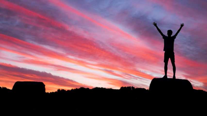 young person standing on a rock with their arms up looking into the sunset. life after cancer image