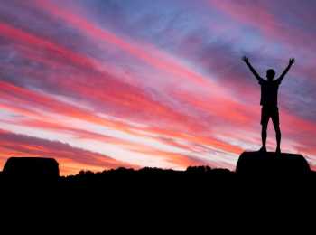 young person standing on a rock with their arms up looking into the sunset. life after cancer image